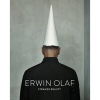 Rückenansicht einer Person in einer schwarzen Jacke, die einen spitzen, weißen Papierhut trägt. Die Person steht vor einer schlichten, grauen Wand. Auf der Jacke steht der Text ERWIN OLAF und darunter STRANGE BEAUTY.