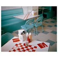 Ein gedeckter Tisch im Vordergrund eines Retro-Diners mit einem rot-weiß karierten Platzdeckchen, Coca-Cola-Servietten und Saucenflaschen. Im Hintergrund sind türkisfarbene Sitzbänke und ein karierter Fliesenboden zu sehen.