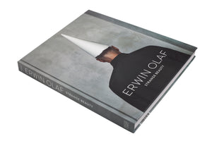Das Fotobuch Erwin Olaf: Strange Beauty liegt schräg auf einem weißen Hintergrund. Der graue Einband zeigt die Rückansicht einer Person mit schwarzem Hemd und einem weißen, kegelförmigen Hut. Der Titel ist in weißer Schrift aufgedruckt.
