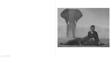 Schwarz-Weiß-Fotografie einer älteren Frau, die auf dem Boden sitzt, während ein Mann seinen Kopf in ihren Schoß legt. Hinter ihnen steht ein großer Elefant in einer staubigen Landschaft.