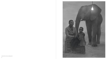 Schwarz-Weiß-Fotografie einer Frau und eines kleinen Kindes in Simbabwe. Die Frau sitzt auf einer Holzkiste, während das Kind neben ihr steht. Direkt hinter ihnen steht ein großer Elefant, über dessen Kopf eine einzelne, leuchtende Glühbirne an einem Kabel hängt.