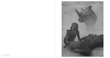 Schwarz-Weiß-Fotografie einer dramatischen Szene: Ein Kind kniet auf staubigem Boden neben einem liegenden Mann und legt eine Hand auf seinen Kopf, während im nebligen Hintergrund ein großes Nashorn steht.