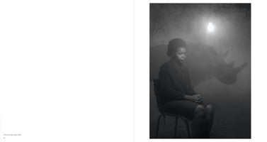 Eine Schwarz-Weiß-Fotografie zeigt eine Frau, die in einer nebligen Nacht auf einem Stuhl sitzt. Sie blickt nachdenklich zu Boden. Hinter ihr taucht die Silhouette eines Nashorns aus dem Dunst auf. Eine einzelne, nackte Glühbirne hängt von oben und beleuchtet die Szene.
