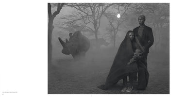 Surreale Schwarz-Weiß-Fotografie einer Familie und zweier Nashörner in einer nebligen Landschaft. Ein Mann steht hinter einer Frau, die ein Baby in einem Tuch hält. Über ihnen leuchtet eine einzelne Glühbirne, die zwischen kahlen Bäumen von oben herabhängt.
