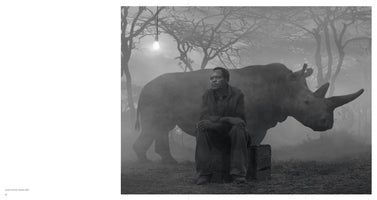 Schwarz-Weiß-Fotografie eines Mannes, der mit nachdenklichem Blick auf einer Holzkiste sitzt. Hinter ihm steht ein großes Nashorn in einer nebligen Landschaft mit kahlen Bäumen. Eine einzelne Glühbirne hängt von oben herab und leuchtet.