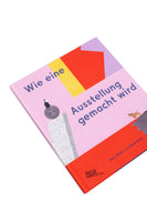 Schrägansicht des Buches Wie eine Ausstellung gemacht wird von Doro Globus und Rose Blake auf einem weißen Hintergrund. Das rosa Cover ist mit bunten, geometrischen Formen und kleinen, verspielten Illustrationen gestaltet, darunter eine Figur auf einer lächelnden Kugel und ein kleiner Hund.