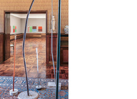 Ein Raum mit gemusterter Tapete, Holzvertäfelung und einem orientalischen Teppich, auf dem moderne, schlanke Skulpturen stehen. Durch eine offene Tür blickt man in einen hellen Ausstellungsraum mit Parkettboden, in dem abstrakte Gemälde mit roten, grünen und blauen Farbblöcken hängen.
