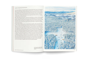 Aufgeschlagenes Buch mit Text auf der linken Seite und einer Luftaufnahme einer Baustelle in einem dichten, schneebedeckten Nadelwald auf der rechten Seite.