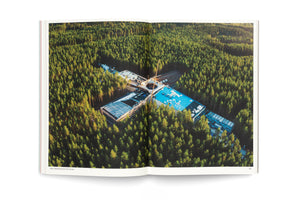 Eine Doppelseite in einem aufgeschlagenen Buch zeigt eine Luftaufnahme eines großen, kreuzförmigen Gebäudes im Bau, das mitten in einem dichten Nadelwald liegt. Die Baumwipfel werden von der tiefstehenden Sonne in goldenes Licht getaucht.