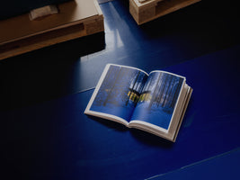Ein aufgeschlagenes Buch liegt auf einer glänzenden, tiefblauen Oberfläche. Auf den Seiten ist ein Foto eines modernen, von innen beleuchteten Hauses in einem verschneiten Wald bei Nacht zu sehen. Im Hintergrund stehen Holzpaletten.