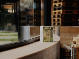 Ein kleines Buch mit dem Titel Making The Plus steht auf einer geschwungenen Fensterbank aus hellem Holz vor einem Fenster. Das Buch spiegelt sich im Glas, und im unscharfen Hintergrund sind gestapelte Holzpaletten in einem industriellen Raum zu sehen.