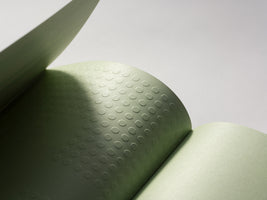 Eine Nahaufnahme zeigt die aufgeschlagenen Seiten eines salbeigrünen Buches. Eine Seite hat eine geprägte Textur aus einem Gitter kleiner Kreise, die durch einen weichen Schattenwurf hervorgehoben wird.