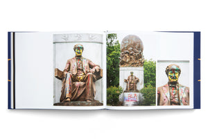 Ein aufgeschlagenes Buch mit blauem Einband liegt auf einem weißen Hintergrund. Auf den Seiten sind drei Fotos einer Bronzestatue eines sitzenden Mannes zu sehen. Die Statue ist mit bunten Farbspritzern beschmiert, und auf dem Sockel des Denkmals sind Graffiti wie BLM und FUCK FACISTS aufgesprüht.