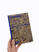 Eine Hand hält ein Buch mit dem Titel Unlimited 023 vor einem weißen Hintergrund. Der Umschlag hat eine Textur aus gepresstem Heu und blauen Blütenblättern. Der Buchrücken und die Schrift sind leuchtend blau.