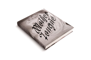Ein graues Hardcover-Buch mit dem Titel Mother Tongue von Camille Henrot liegt schräg auf einer weißen Oberfläche. Der Titel ist in einer großen schwarzen Schreibschrift über ein Schwarz-Weiß-Foto gedruckt.