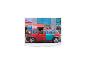 Ein aufgeschlagenes Buch zeigt das Foto eines zweifarbigen Autos in Berlin. Die vordere rote Hälfte des Wagens passt zur roten Markise eines Cafés, die hintere türkise Hälfte zur Fassade des Nachbargebäudes.