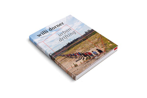 Das Buch urban drifting von Willi Dorner, schräg auf einer weißen Fläche liegend. Das Cover zeigt eine lange Reihe von Menschen auf einem sandigen Hang unter blauem Himmel, überlagert von dünnen roten Linien, die eine geometrische Form bilden.