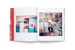Ein aufgeschlagenes Buch, das zwei künstlerische Fotografien zeigt. Auf dem linken Foto liegen ein Mann und eine Frau in roten Hosen waagerecht auf Küchenschränken. Auf dem rechten Foto balanciert eine Person in einer vollgestellten Küche auf der Arbeitsplatte.