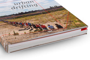 Ein dickes Buch mit dem Titel urban drifting liegt auf einer weißen Oberfläche. Das Coverfoto zeigt eine lange Menschenschlange von hinten, die durch eine karge, offene Landschaft geht. Rote grafische Linien sind über das Bild gelegt.