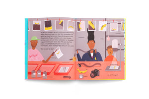 Eine illustrierte Doppelseite aus einem Kinderbuch, die den Prozess der analogen Fotografie in einer Dunkelkammer zeigt. Drei Personen entwickeln Fotos von einer Banane. Verschiedene Geräte wie Kamera, Film, Chemikalien und ein Vergrößerungsgerät sind mit deutschen Begriffen beschriftet. Die fertigen Fotos hängen zum Trocknen an einer Leine.