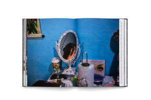 Aufgeschlagenes Buch mit einem Foto, das die Reflexion eines teilweise verschatteten Gesichts in einem kleinen Spiegel zeigt. Der Spiegel steht auf einem Schminktisch mit Kosmetika vor einer blauen Wand.