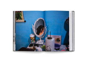 Ein aufgeschlagenes Buch, das eine Fotografie über eine Doppelseite zeigt. In einem verzierten Schminkspiegel auf einem unordentlichen Tisch reflektiert sich das schwach beleuchtete Gesicht einer Person. Der Hintergrund ist eine leuchtend blaue Wand, die einen intimen und melancholischen Eindruck erweckt.