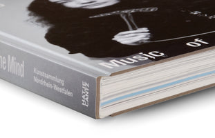 Detailansicht der oberen Ecke eines Buches mit grauem Buchrücken und einem Schwarz-Weiß-Foto auf dem Cover. Sichtbar sind die Schriftzüge Music of, Kunstsammlung Nordrhein-Westfalen und Hatje Cantz. Die Seitenkante des Buches weist einen hellblauen Streifen auf.