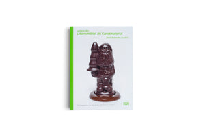 Das Buchcover von Lexikon der Lebensmittel als Kunstmaterial zeigt eine Schokoladenfigur in Form eines Weihnachtsmanns, dessen Mütze wie ein Buttplug geformt ist. Das Buch mit leuchtend grünem Buchrücken steht aufrecht vor weißem Hintergrund.