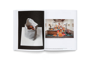 Ein aufgeschlagenes Kunstbuch auf einer weißen Oberfläche. Links ist die Fotografie einer Skulptur, die eine Papiertüte mit Donuts zeigt. Rechts ist die Fotografie einer raumfüllenden Installation in einer Galerie mit Maschinen und großen Haufen orangefarbener, donutähnlicher Objekte.