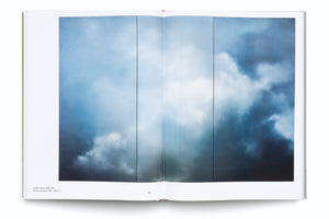 Ein aufgeschlagenes Kunstbuch zeigt auf einer Doppelseite eine Reproduktion des Gemäldes Wolken (blau) von 1970. Das Bild eines Himmels mit dramatischen, weißen und dunkelblauen Wolken ist in vier vertikale Felder unterteilt.