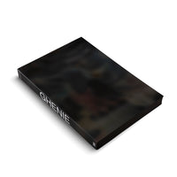Ein schwarzes, gebundenes Kunstbuch mit dem Titel GHENIE in weißer Schrift auf dem Buchrücken, das schräg auf einer weißen Fläche liegt. Der Bucheinband zeigt ein dunkles, abstraktes Gemälde.