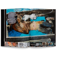 Ein aufgeschlagenes Kunstbuch zeigt ein expressionistisches Gemälde über eine Doppelseite. Eine liegende, entstellte Figur mit einem grauen, helmartigen Kopf ist auf einem leuchtend blauen Hintergrund zu sehen, umgeben von dunklen, kräftigen Pinselstrichen. Am unteren Rand sind kleinere Skizzen und der Titel RESURRECTION 1 abgebildet.