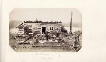Eine alte Sepia-Fotografie zeigt drei Frauen und einen Mann, die als Arbeiter posieren, vor einer rustikalen Blockhütte an einem See. Die Umgebung ist eine gerodete Lichtung mit zahlreichen Baumstümpfen und einer großen Säge, die im Vordergrund an einem Stumpf lehnt.