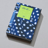 Das Buch alia ali ... von Hatje Cantz liegt auf einem grauen Untergrund. Es hat einen Stoffeinband, der vorne dunkelblau mit weißen Tupfen und am Buchrücken mit einem blau-weiß-schwarzen grafischen Muster versehen ist. Ein lindgrünes Etikett auf dem Cover zeigt den Buchtitel.