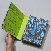 Eine Hand hält ein aufgeschlagenes Buch mit einem blau-braunen Batik-Einband. Die Innenseite ist leuchtend grün und mit blauem Text bedruckt, der die Worte Neeli fruit, Bunni clouds und Surrounded by Noor zeigt.