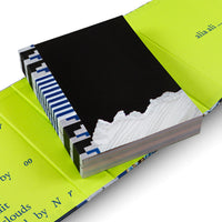 Ein kunstvoll gestaltetes Buch liegt in seinem offenen, neongrünen Schuber. Der freiliegende Buchrücken hat ein blau-weiß-schwarzes Textilmuster. Der schwarze Buchdeckel ist unten abgerissen und enthüllt eine weiße, strukturierte Oberfläche, die an Schnee oder Stoff erinnert.