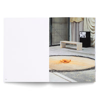 Aufgeschlagenes Kunstbuch. Die linke Seite ist leer, nur mit der Seitenzahl 90. Rechts ist eine Kunstinstallation in einer Halle zu sehen: eine kreisrunde Bodenarbeit mit orangem Pigment, dahinter eine schlichte, helle Bank und ein schwarzer Kasten mit einem Monitor, der ein Schwarz-Weiß-Bild zeigt.