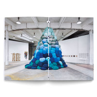 Aufgeschlagene Buchdoppelseite, die eine große, pyramidenförmige Skulptur in einer Kunstgalerie zeigt. Die Skulptur besteht aus vielen runden, weichen Kissen in verschiedenen Blautönen, die von dunkel am Boden zu hell an der Spitze verlaufen. Im Hintergrund des industriell anmutenden Raums sind weitere Kunstwerke an den weißen Wänden zu sehen.