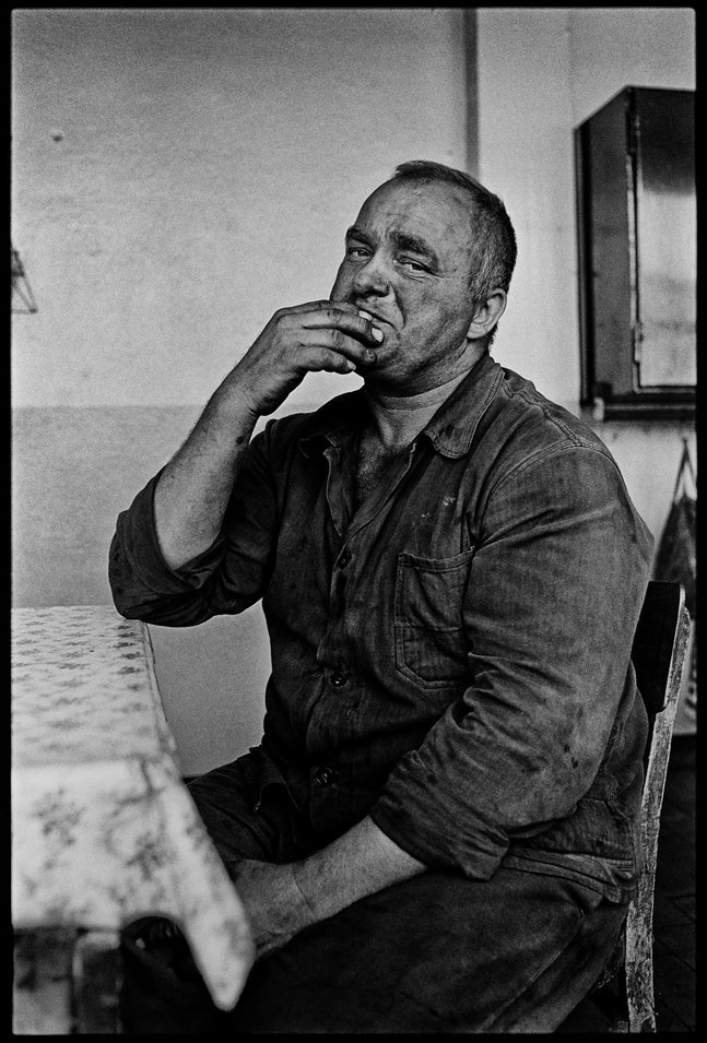 Eine körnige Schwarz-Weiß-Fotografie eines Arbeiters, der an einem Tisch sitzt. Er hat ein schmutziges Gesicht und schmutzige Arbeitskleidung an, führt eine Zigarette zum Mund und blickt mit einem nachdenklichen Ausdruck direkt in die Kamera.