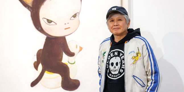 Der japanische Künstler Yoshitomo Nara steht neben einem seiner Gemälde. Er trägt eine Baseballkappe und eine bestickte Bomberjacke über einem schwarzen Hoodie. Das Kunstwerk zeigt eine seiner charakteristischen, mürrisch blickenden Kinderfiguren in einem Tierkostüm, die auf einer Spielzeugente sitzt.