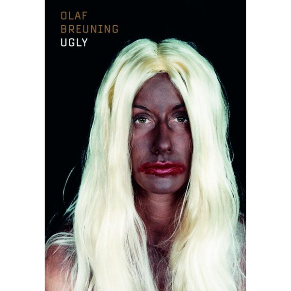Eine Person mit langer, blonder Perücke, dunkel bemaltem Gesicht und verschmiertem roten Lippenstift auf schwarzem Hintergrund.