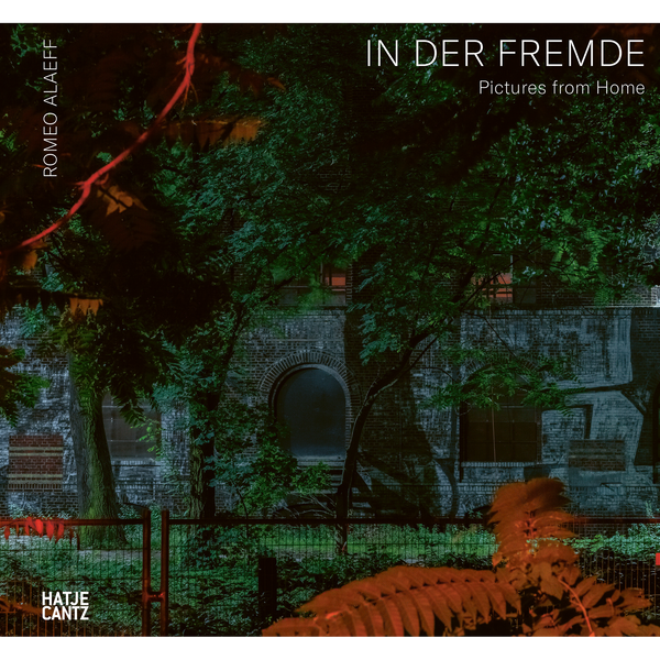 Buchcover für Romeo Alaeffs In der Fremde. Eine nächtliche Aufnahme eines Backsteingebäudes, gesehen durch einen Zaun und dichte, grüne Bäume, mit roten Lichtakzenten auf den Blättern im Vordergrund.