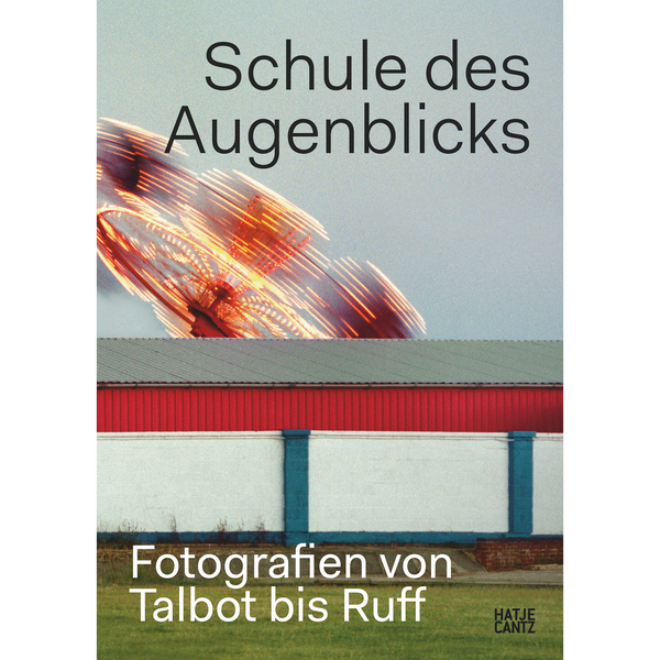 Das Buchcover zu Schule des Augenblicks zeigt ein zweigeteiltes Bild: oben ein beleuchtetes Fahrgeschäft mit Bewegungsunschärfe, unten die rot-weiß-blaue Wand eines Gebäudes.