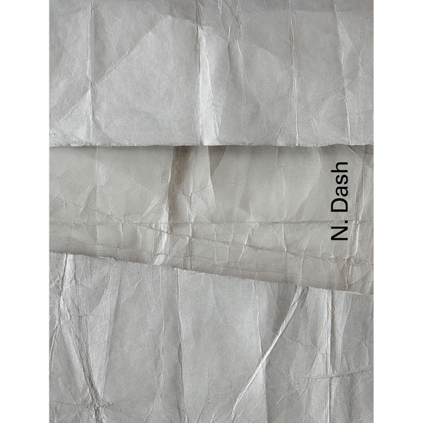 Eine Nahaufnahme von zerknittertem, grauem Papier in überlappenden Schichten. Am rechten Rand steht vertikal der Name N. Dash.