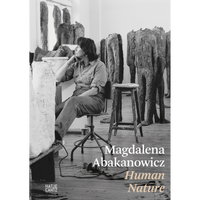 Eine Schwarz-Weiß-Fotografie der Künstlerin Magdalena Abakanowicz, die nachdenklich in ihrem Atelier sitzt, umgeben von ihren großen, kopflosen Skulpturen.