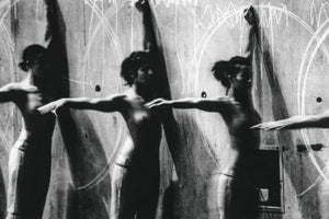 Körniges Schwarz-Weiß-Foto, das eine Bewegungssequenz von drei Personen mit nacktem Oberkörper zeigt, die vor einer rauen Wand mit weißen Kreidezeichnungen von Kreisen und Linien stehen. Ihre Arme sind in dynamischen Posen ausgestreckt und ihre Körper durch die Bewegung unscharf.