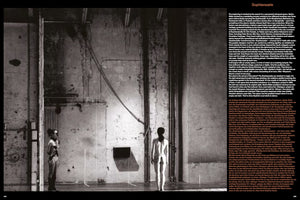Schwarz-Weiß-Fotografie einer Performance in einem kargen Industrieraum. Eine Frau steht mit dem Rücken zur Kamera im Zentrum, während ein Mann links im Profil steht. Die Wände sind roh und fleckig, und ein Kabel hängt von der Decke.