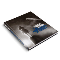 Ein schwarzes Hardcover-Buch mit dem Titel Robert Longo: The Acceleration of History liegt auf einer weißen Oberfläche. Das Cover zeigt eine dramatische Schwarz-Weiß-Aufnahme einer Reiterstatue in Silhouette, die von unten aufgenommen und von Rauch oder Nebel umgeben ist. Der Titel ist in weißen und blauen Buchstaben auf dem Cover und dem Buchrücken abgebildet.