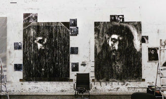 Ein Künstleratelier mit einer schmutzigen weißen Wand, an der zwei große, düstere Porträts in Schwarz-Weiß hängen. Die Gesichter auf den Leinwänden sind durch viele vertikale schwarze Linien teilweise verdeckt. Kleinere Skizzen sind um die großen Bilder herum angebracht. In der Mitte steht ein schwarzer Bürostuhl auf einem Holzboden.