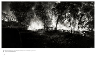 Eine dramatische Schwarz-Weiß-Aufnahme eines Waldbrandes bei Nacht. Ein einzelnes Auto fährt auf einer Straße, während rechts davon Bäume und im Hintergrund ein Hügel in hellen Flammen stehen und dicker Rauch aufsteigt.